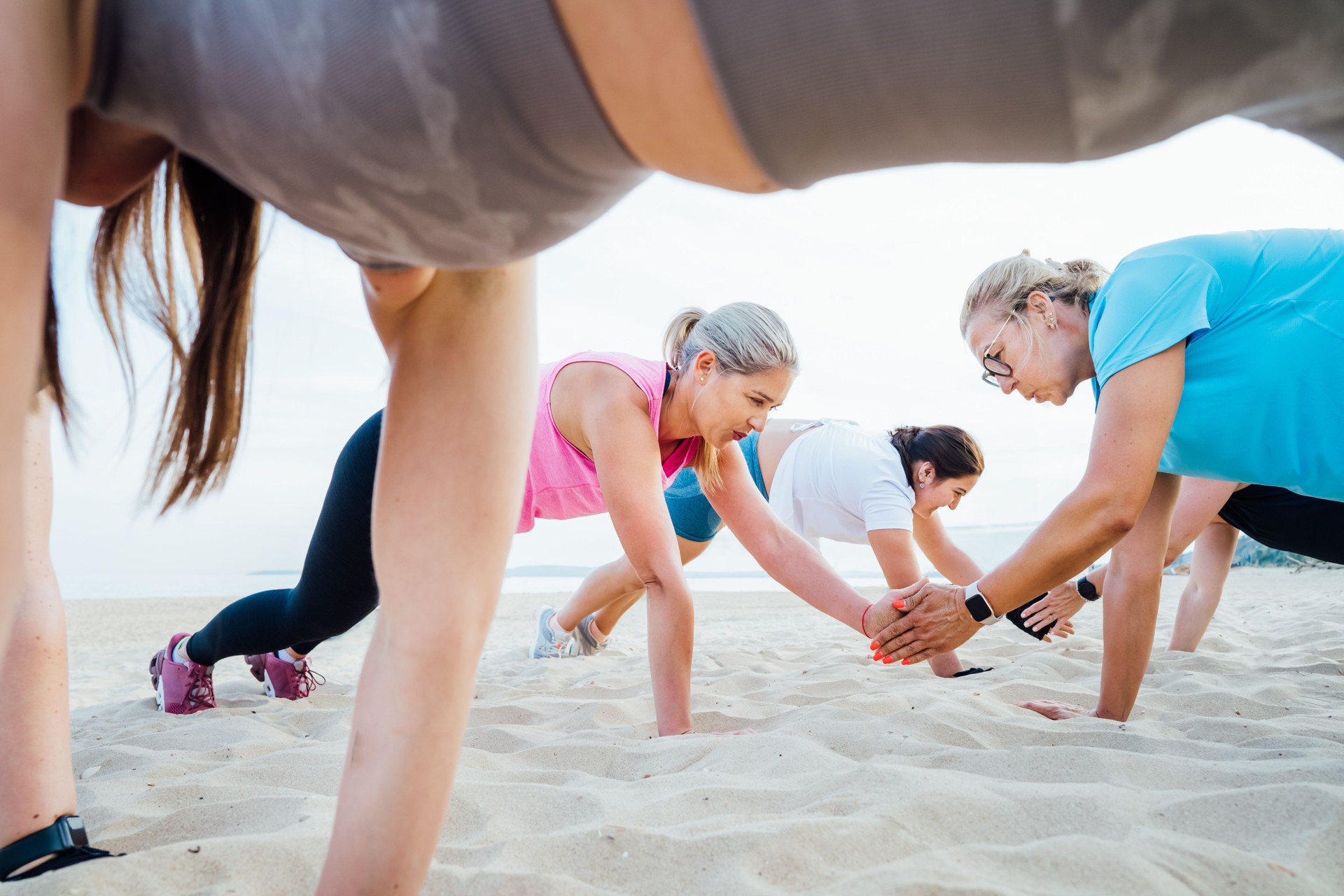 Women of various ages doing fitness workouts in class exercise with coach on beach. Ladies doing paired plank exercises and high-fiving each other. Sport for health and wellbeing. Active lifestyle